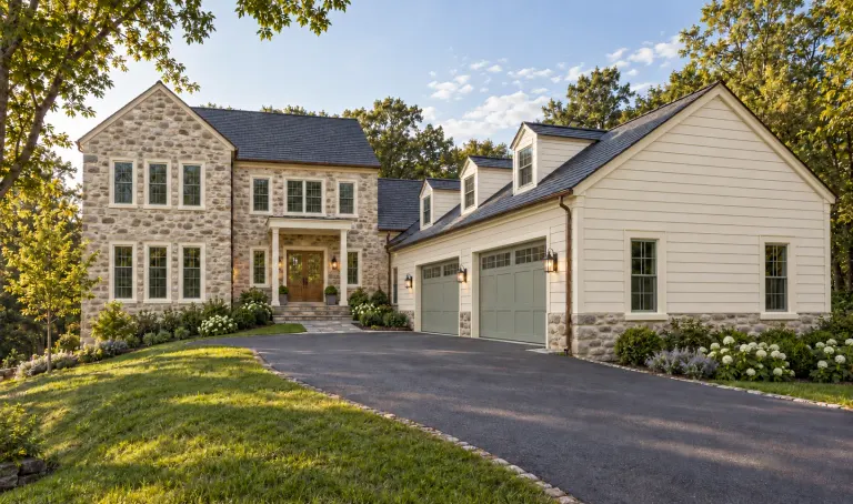 Stone and white house with tall windows, dormers, an attached garage and landscaped driveway.