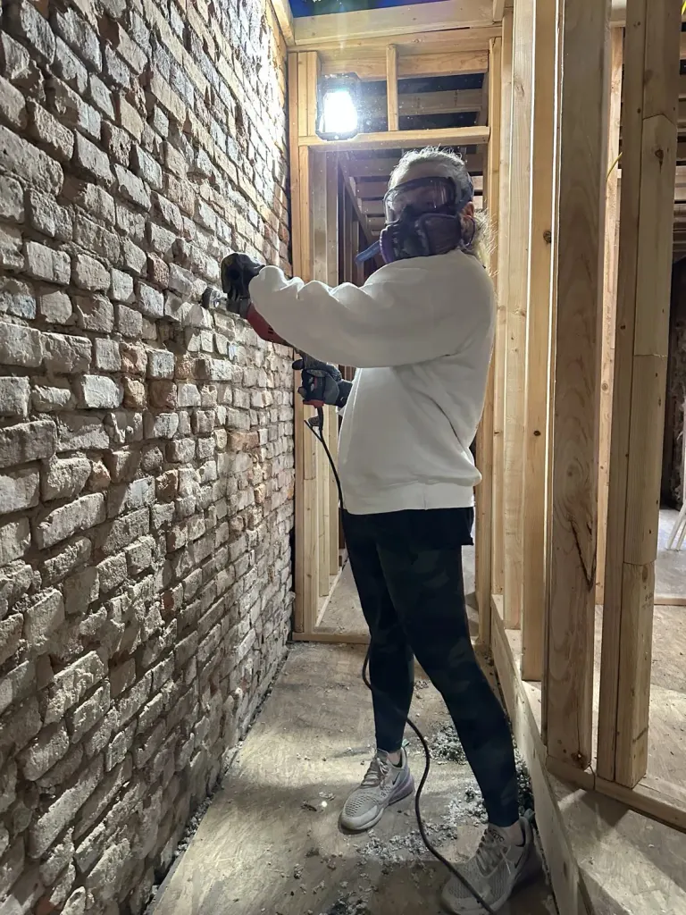 A worker wearing protective gear while cleaning exposed brick beside open framing.