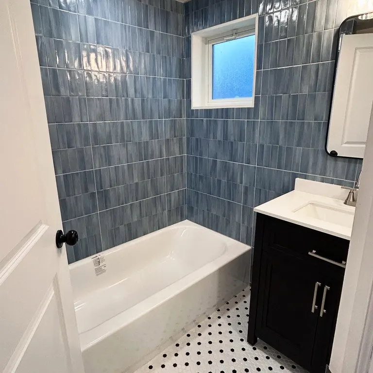 Bathroom with blue-gray tile, white bathtub, black hardware and light window.