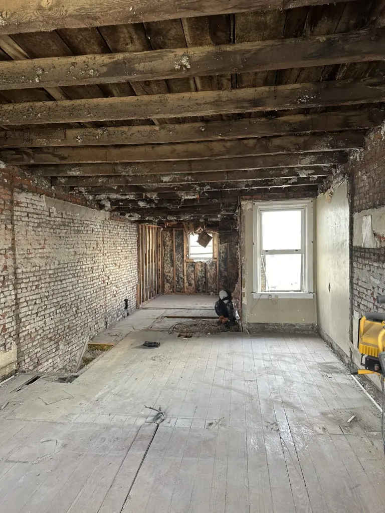 A room during demolition, with brick walls, bare joists, and a stripped floor.